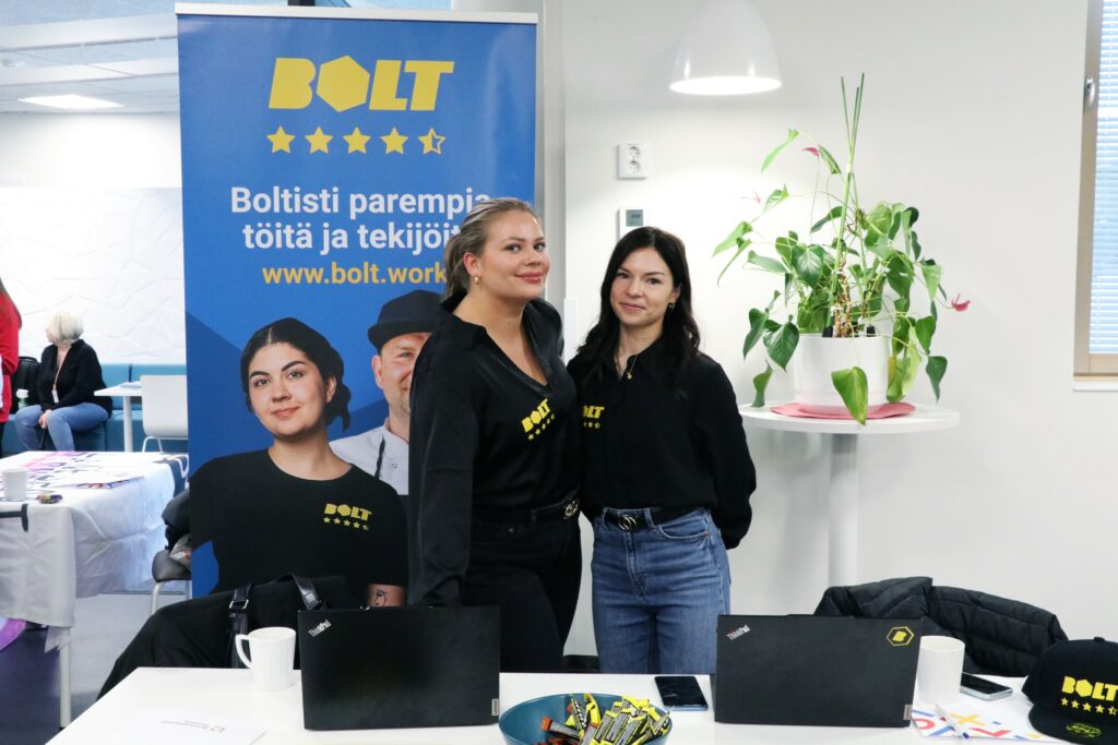 Kaksi naista poseeraamassa kameraan. Yllään heillä on Bolt-yrityksen paidat, ja taustalla näkyy myös Boltin roll up -juliste.